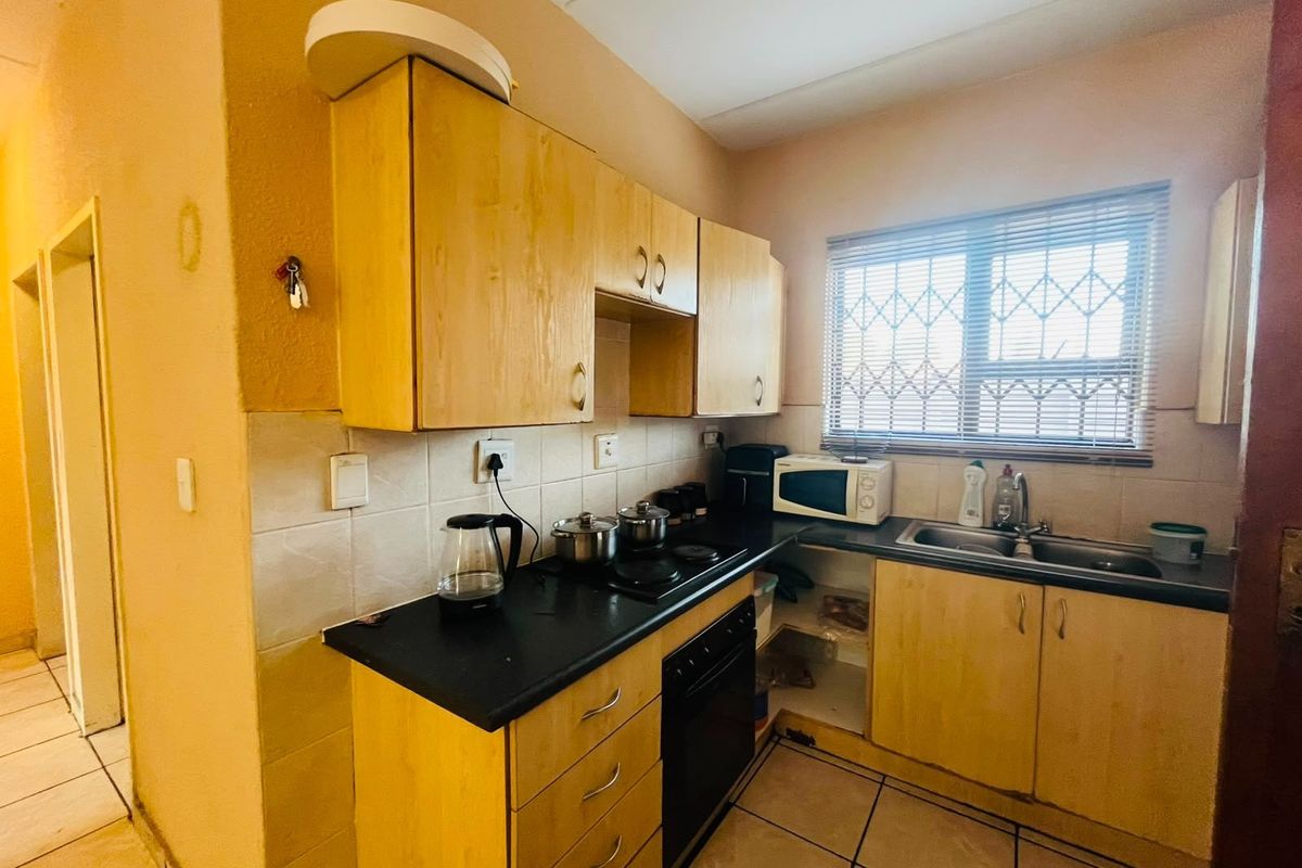 A compact kitchen featuring light wood-tone cabinetry, dark laminate countertops, a built-in oven and hob, and tiled flooring with security bars on the window.