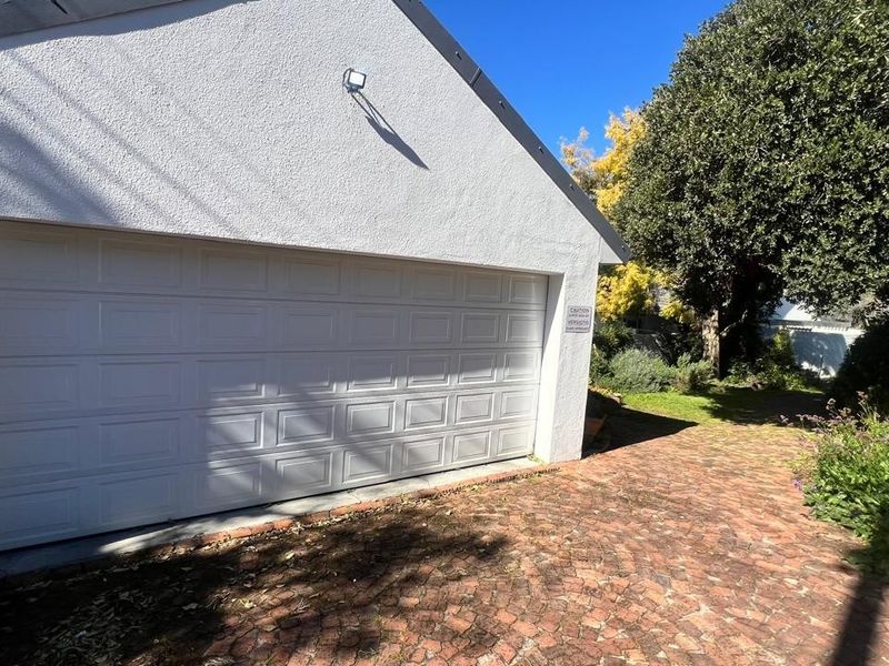 A detached garage featuring a white sectional door, stucco exterior finish, and a brick-paved driveway surrounded by mature garden landscaping.