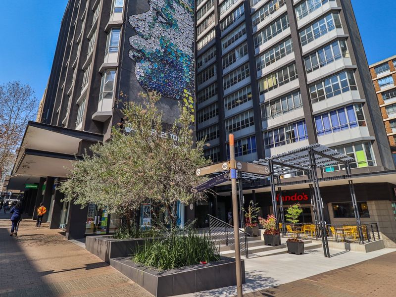 Exterior view of a multi-story commercial building featuring a prominent wall mural, paved pedestrian plaza, and integrated outdoor seating area with a metal pergola.