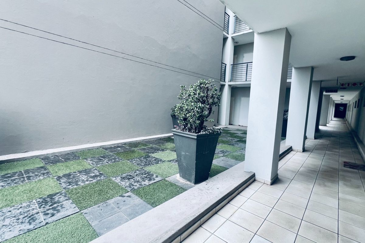 A covered outdoor walkway and courtyard area featuring a checkered pattern of artificial grass and stone pavers with structural concrete pillars.