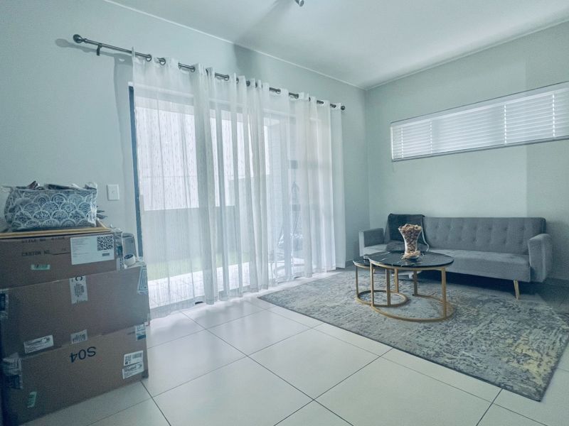 A bright living room featuring large sliding glass doors with sheer curtains, tiled flooring, and horizontal window blinds.
