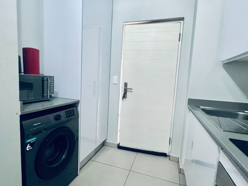 A compact, modern scullery featuring sleek white cabinetry, stone countertops, a stainless steel sink, and dedicated space for laundry appliances.