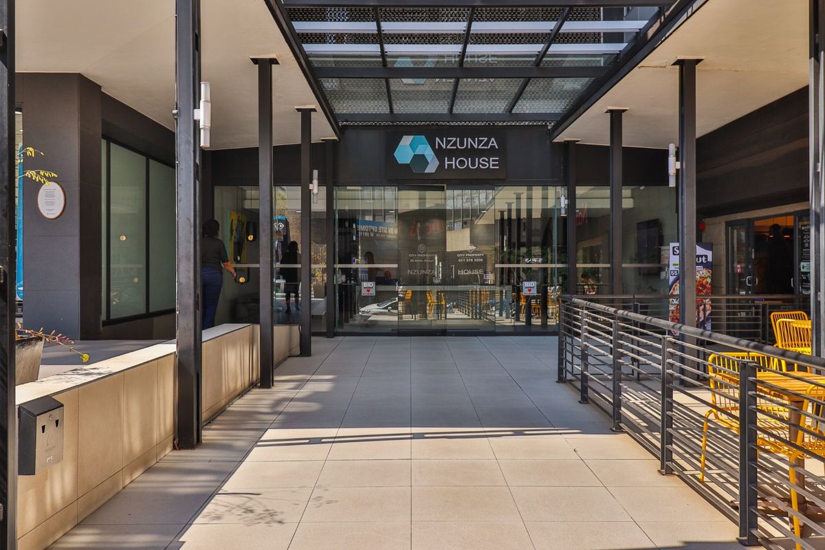 Modern commercial building entrance featuring a tiled walkway, glass facade, and a protective metal-framed canopy.