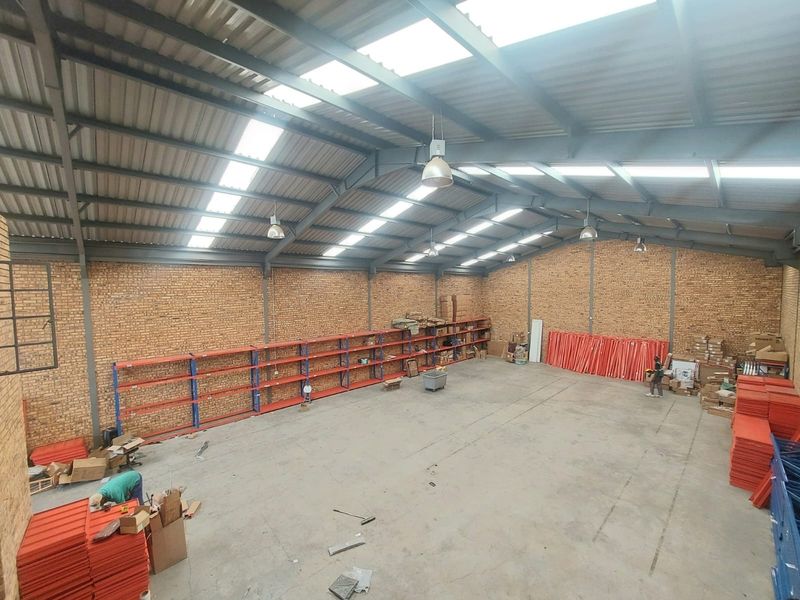 Spacious industrial warehouse featuring high ceilings with exposed steel trusses, abundant natural light from skylights, facebrick walls, and extensive built-in industrial shelving.