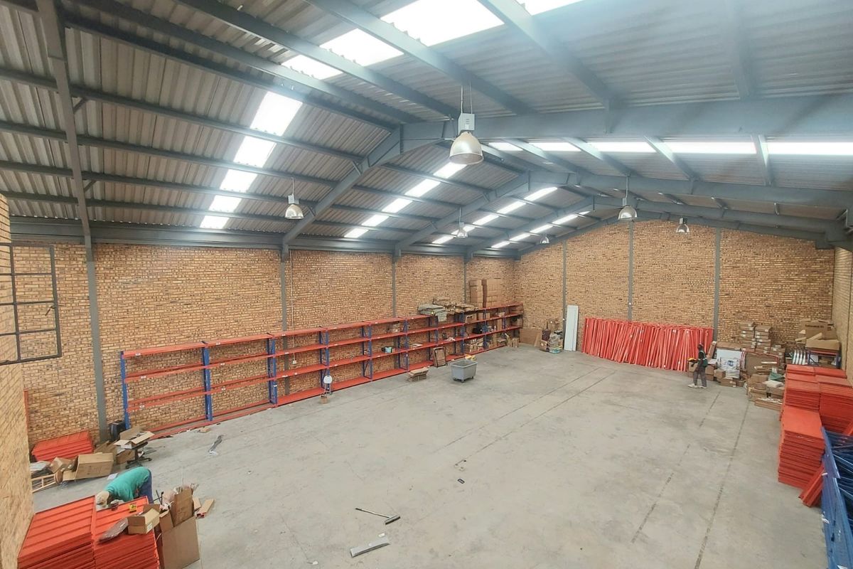 Spacious industrial warehouse featuring high ceilings with exposed steel trusses, abundant natural light from skylights, facebrick walls, and extensive built-in industrial shelving.