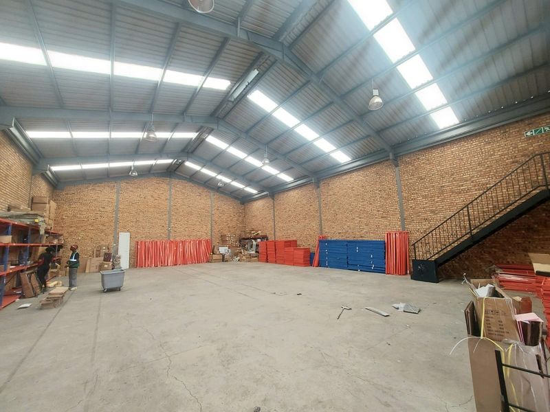 Spacious industrial warehouse featuring exposed brick walls, high-clearance steel portal frame roof with natural lighting, and a metal staircase leading to an upper level.