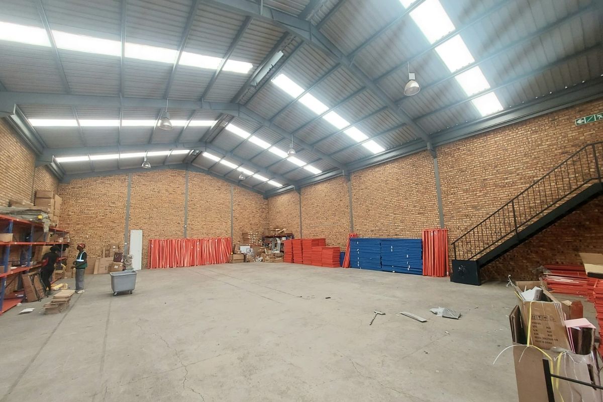 Spacious industrial warehouse featuring exposed brick walls, high-clearance steel portal frame roof with natural lighting, and a metal staircase leading to an upper level.