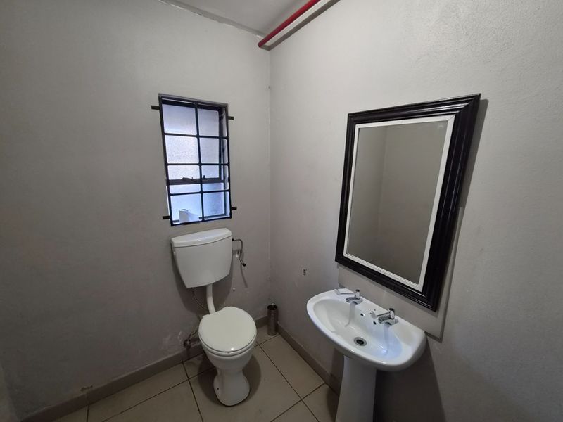 A functional industrial-style bathroom featuring a pedestal basin, toilet, and security-barred window, requiring minor cosmetic updates to improve its overall appearance.