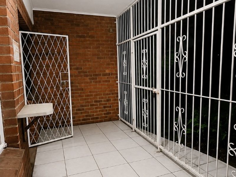 A covered patio or balcony area featuring tiled flooring, exposed brick walls, and secure metal security gates.