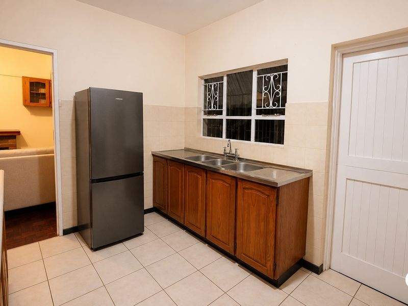 A functional kitchen featuring wooden cabinetry, a stainless steel double sink, tiled splashbacks, and tiled flooring, with direct access to the living area.