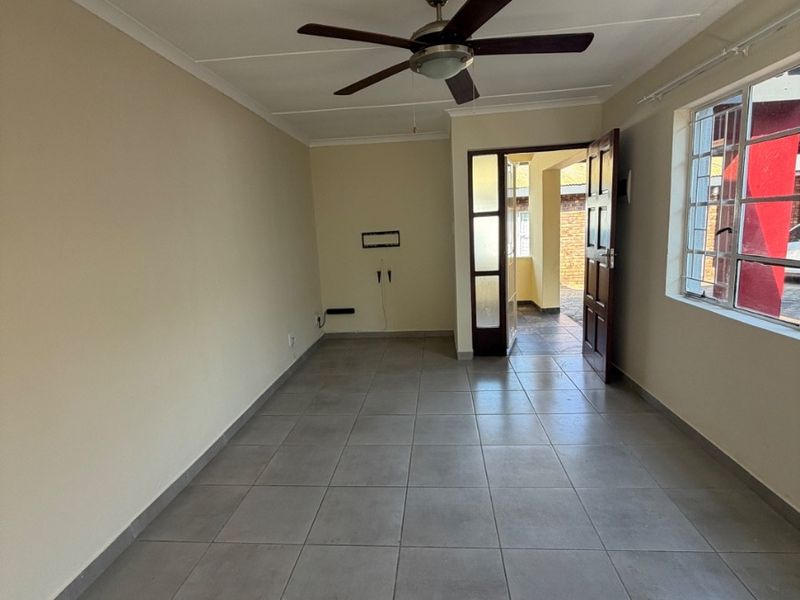 An empty living room featuring neutral tiled flooring, a ceiling fan, and direct access to an outdoor area through a timber door.
