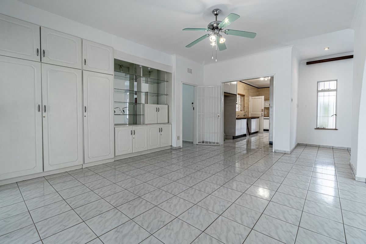 Spacious living area featuring neutral tiled flooring, built-in storage cabinetry with glass display shelving, and a ceiling fan, with direct access to the kitchen.