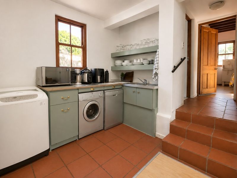 A functional laundry area featuring built-in cabinetry, open shelving, a stainless steel sink, and terracotta tiled flooring, with stairs leading to an adjacent room.