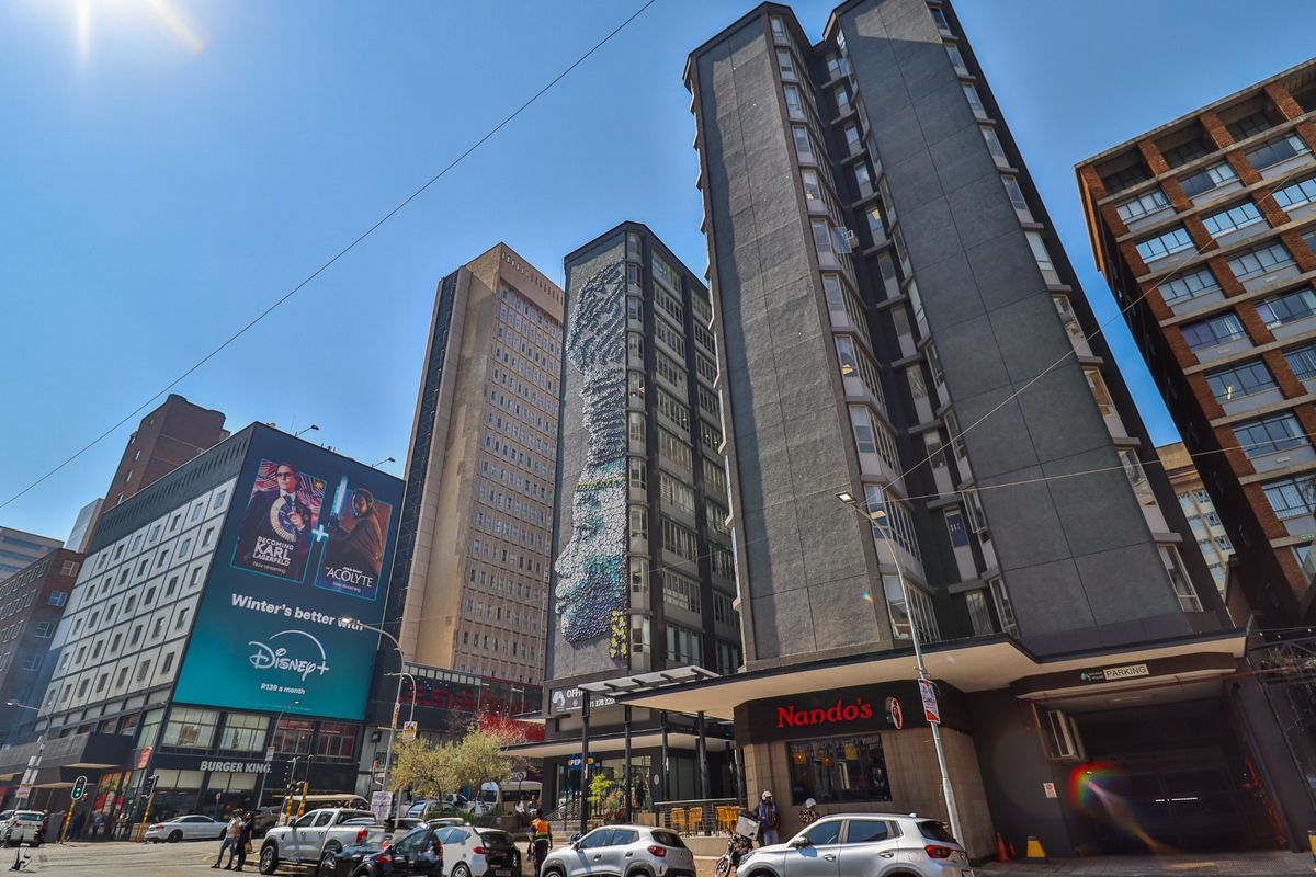 A street-level view of a commercial urban block featuring high-rise buildings, retail storefronts, and large-scale advertising signage in a busy city environment.