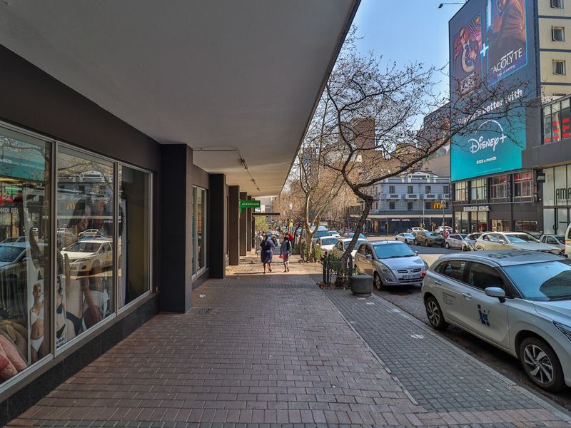 A view of a commercial street sidewalk with covered walkways and storefront display windows in an urban setting.