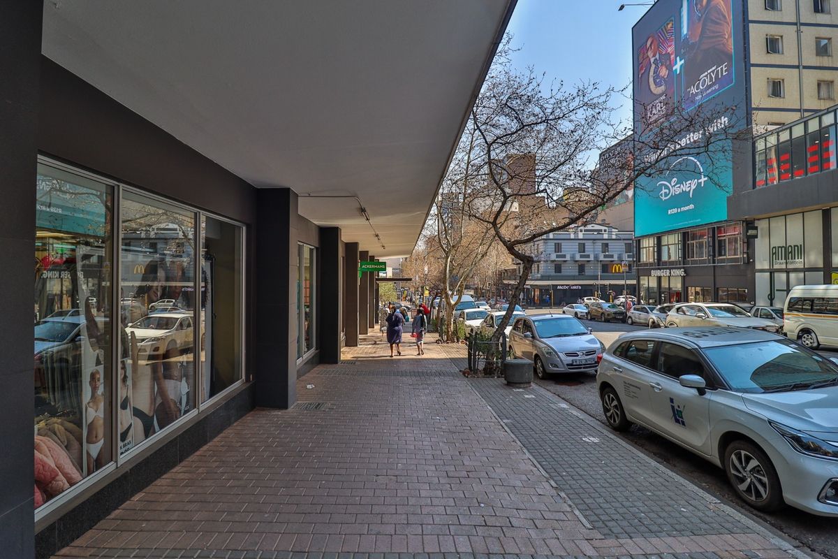 A view of a commercial street sidewalk with covered walkways and storefront display windows in an urban setting.