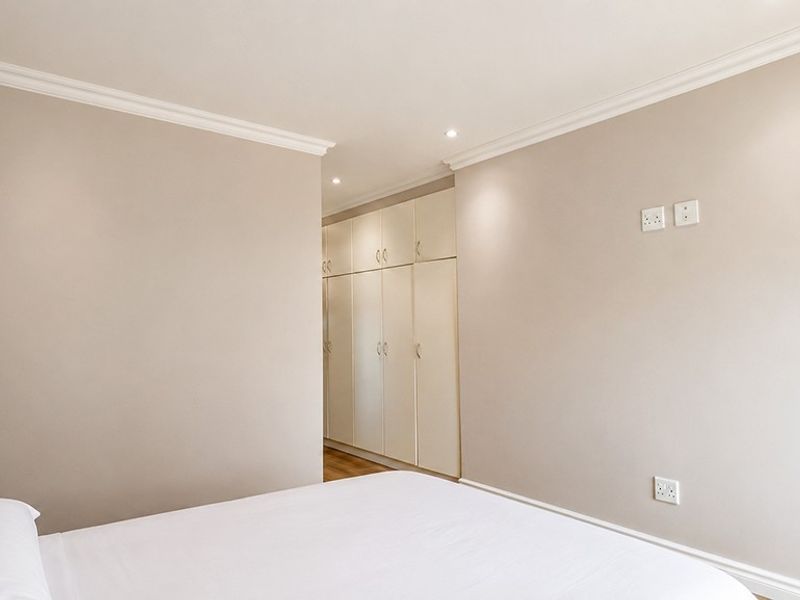 A clean, neutral bedroom featuring built-in cupboards, recessed downlighting, and decorative coving, providing a bright and well-maintained living space.