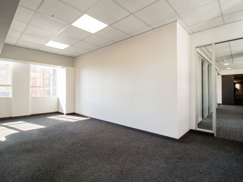 A bright, empty commercial office space featuring a suspended ceiling, recessed lighting, carpeted flooring, and a glass partition wall.