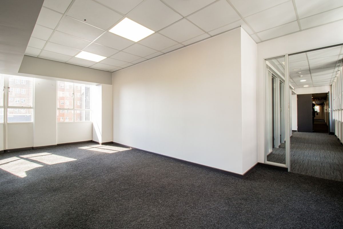 A bright, empty commercial office space featuring a suspended ceiling, recessed lighting, carpeted flooring, and a glass partition wall.