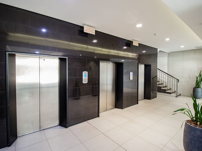 Modern commercial building lobby featuring a bank of three stainless steel elevators, black tiled feature wall, and access to a staircase.