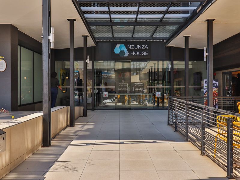 Modern commercial building entrance featuring a tiled walkway, glass facade, and a protective metal-framed canopy.