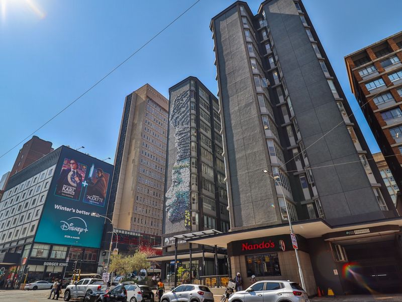 A street-level view of a commercial urban block featuring high-rise buildings, retail storefronts, and large-scale advertising signage in a busy city environment.