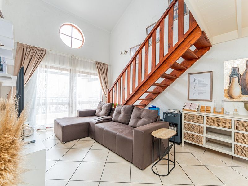 A bright living room featuring a wooden staircase, vaulted ceilings, tiled flooring, and a circular window with access to an outdoor balcony.
