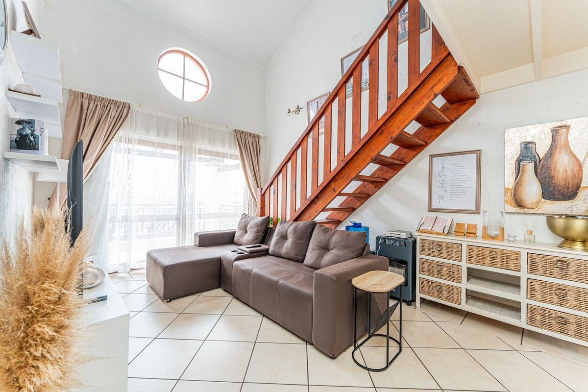 A bright living room featuring a wooden staircase, vaulted ceilings, tiled flooring, and a circular window with access to an outdoor balcony.
