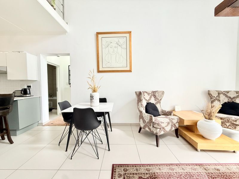 An open-plan living and dining area featuring tiled floors, a compact kitchen, and a mezzanine level with a modern staircase railing.