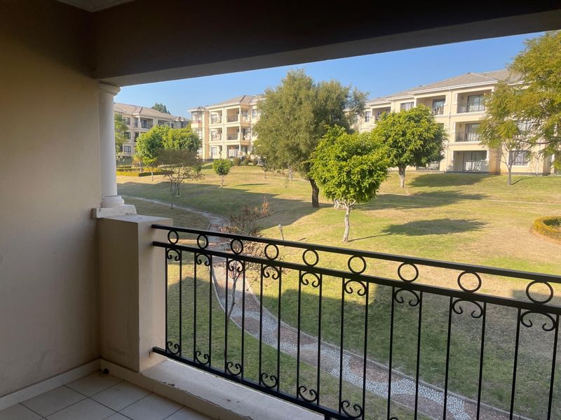 A covered balcony featuring a wrought iron balustrade and tiled flooring, overlooking a landscaped communal garden area within the apartment complex.