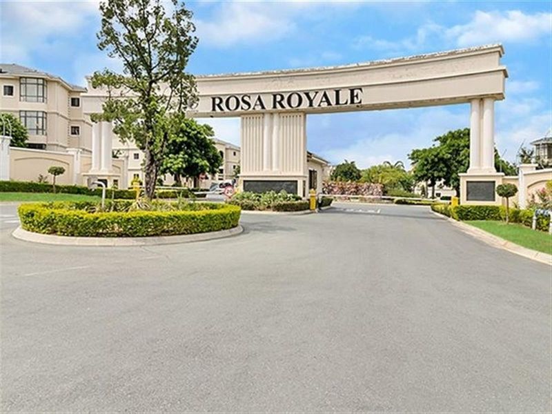 The main entrance to the Rosa Royale apartment complex, featuring a grand architectural archway, security boom gate, and well-maintained landscaped surroundings.