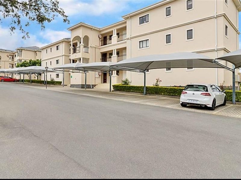 Exterior view of a multi-story apartment complex featuring paved parking bays with shade cloth carports and well-maintained landscaped hedging.