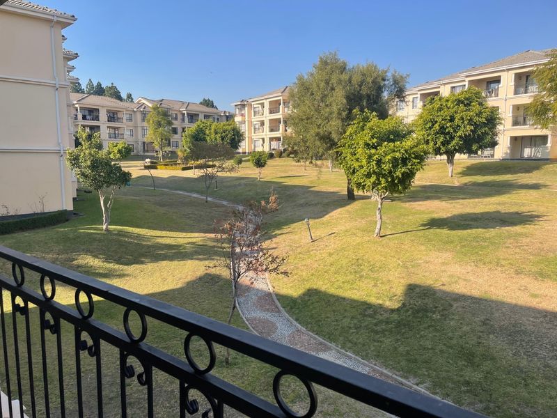 View of the apartment complex communal garden area featuring manicured lawns, mature trees, and paved pathways, viewed from a private balcony.