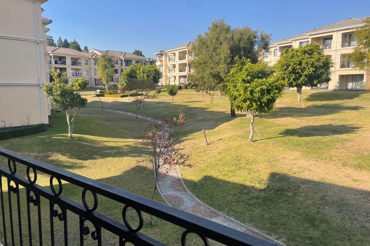 View of the apartment complex communal garden area featuring manicured lawns, mature trees, and paved pathways, viewed from a private balcony.