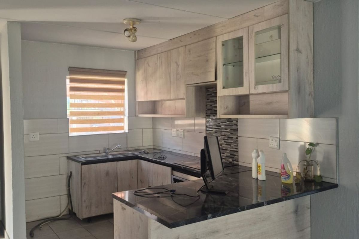 A functional kitchen featuring light wood-look cabinetry, dark laminate countertops, tiled backsplash, and neutral floor tiles in an open-plan layout.