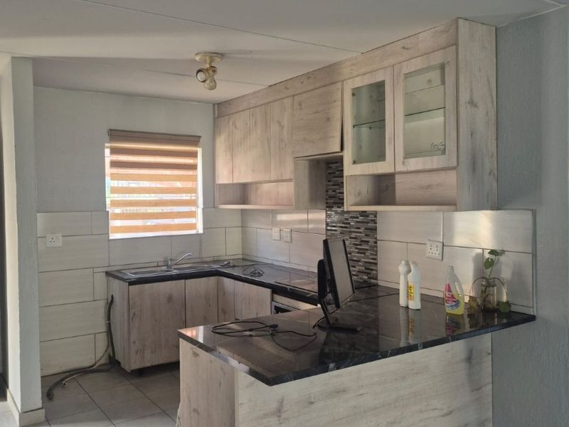 A functional kitchen featuring light wood-look cabinetry, dark laminate countertops, tiled backsplash, and neutral floor tiles in an open-plan layout.