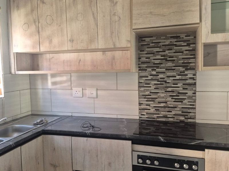 A functional kitchen featuring light wood-look cabinetry, dark countertops, a mosaic tile backsplash, and integrated oven and hob.