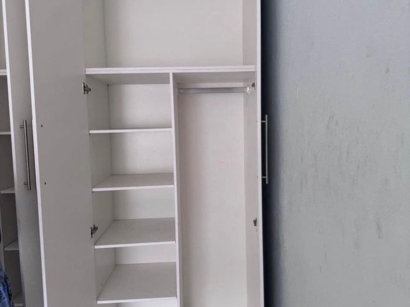 A white built-in wardrobe featuring internal shelving and a hanging rail, set against a plain wall with tiled flooring.