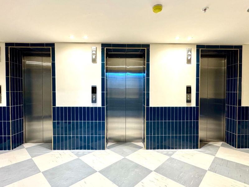 A modern elevator lobby featuring three stainless steel lift doors, accented by blue ceramic wall tiles and a classic checkered floor pattern.