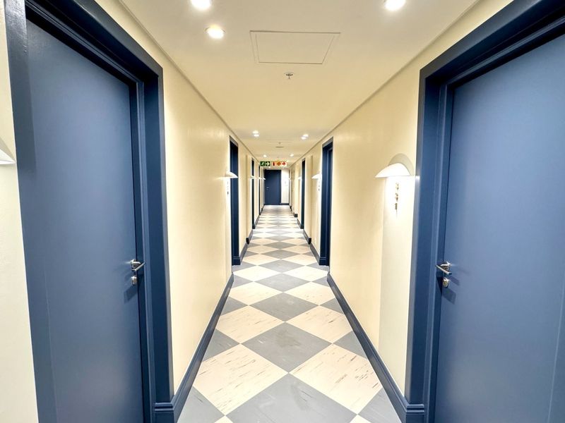 A well-maintained interior hallway featuring a distinctive checkered floor pattern, blue painted doors, and modern recessed and wall-mounted lighting.