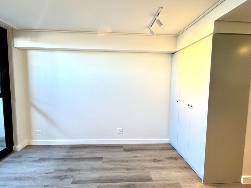 A bright, modern bedroom featuring neutral walls, laminate flooring, built-in cupboards, and a sliding glass door leading to an outdoor area.