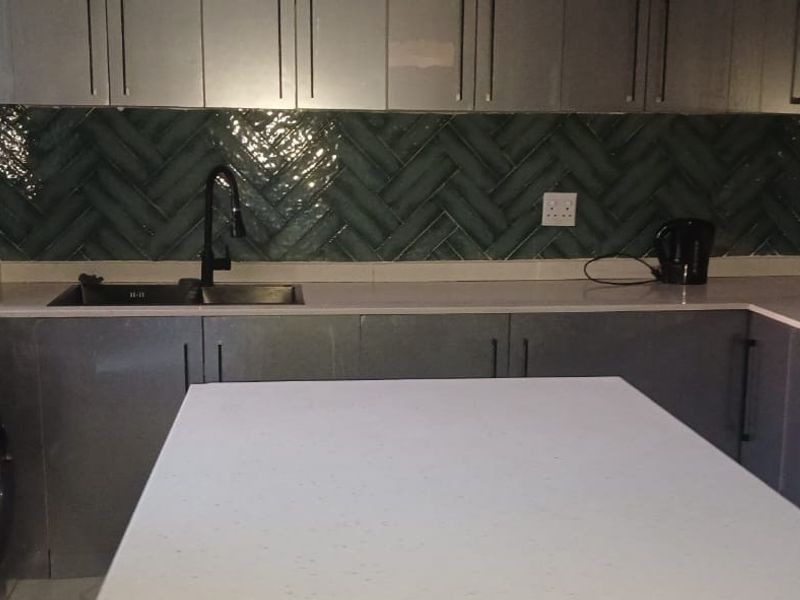Modern kitchen featuring sleek grey cabinetry, stone countertops, and a stylish green herringbone tile backsplash with integrated appliances.