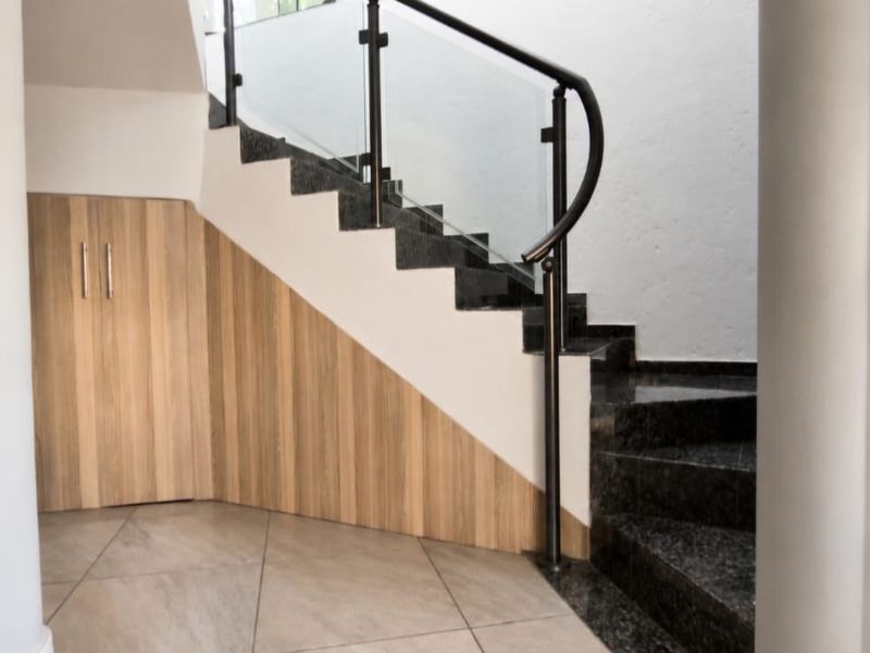 A modern hallway featuring a staircase with a glass balustrade and integrated under-stair storage cabinetry, finished with neutral floor tiles.