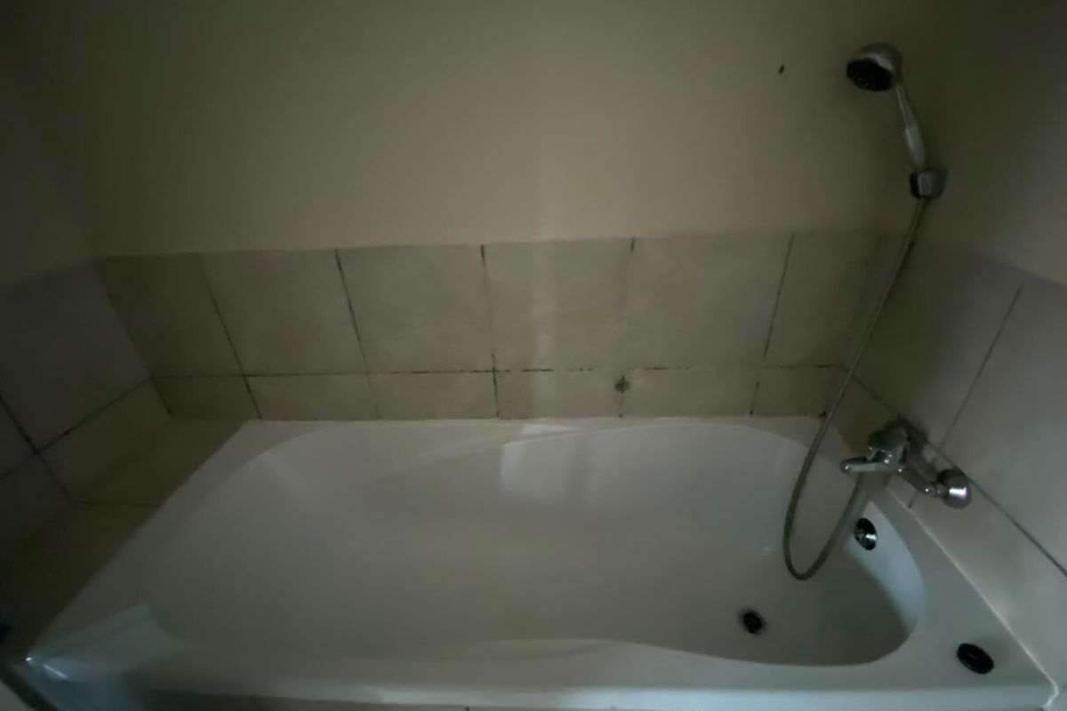 A basic bathroom featuring a built-in bathtub with a handheld shower fixture and tiled splashback, requiring some maintenance and cosmetic updates.