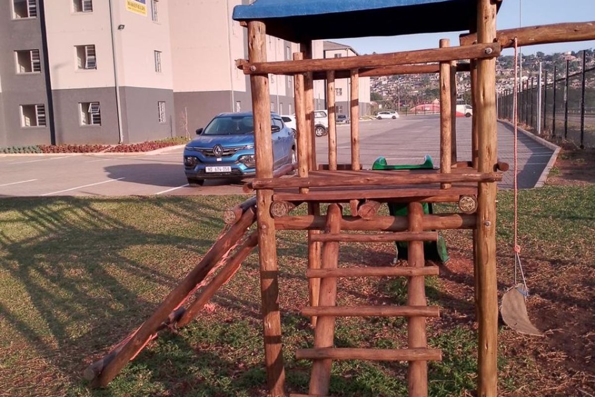 Exterior view of an apartment complex featuring a communal playground area with wooden climbing structure and adjacent paved parking.