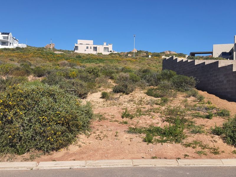 A vacant residential plot featuring a sloped landscape, natural vegetation, and an existing concrete block retaining wall, situated in a developed neighborhood.