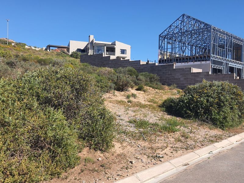 A vacant residential plot featuring a partially constructed steel frame building and a concrete retaining wall on a sloped landscape.