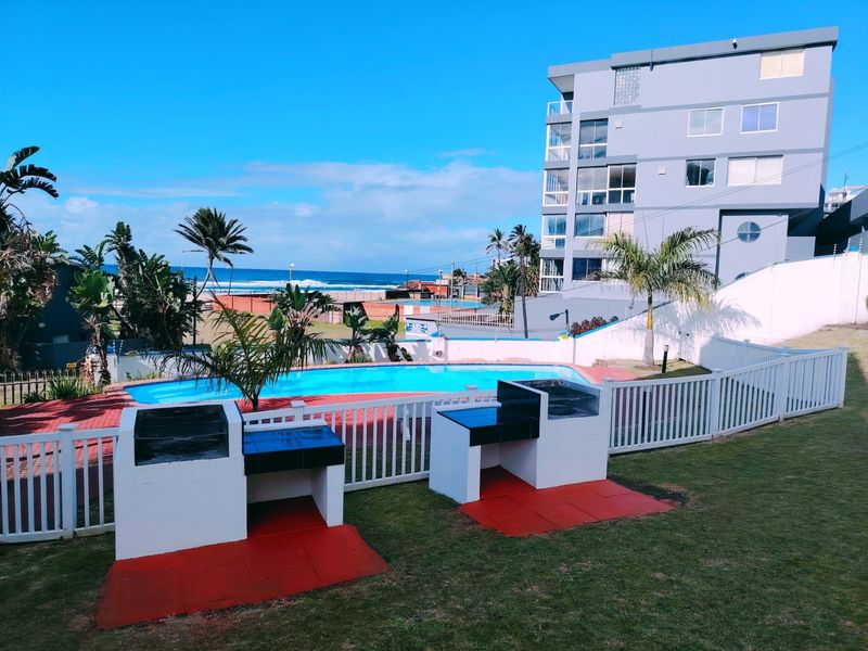 Communal pool area featuring built-in braai stations, paved surrounds, and landscaped garden, offering views of the nearby coastline and ocean.