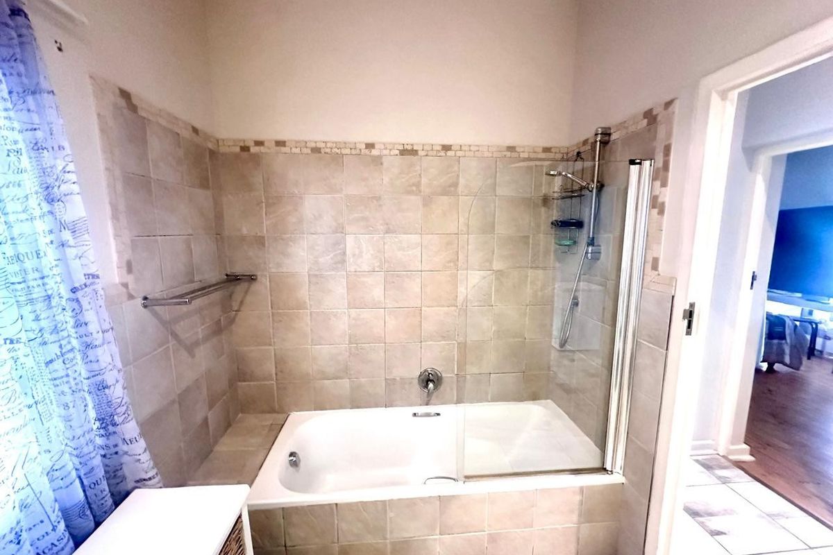 A functional bathroom featuring a built-in bathtub with a glass shower screen and neutral wall tiling.