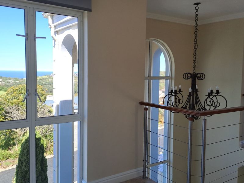 A bright, open hallway featuring a decorative wrought iron chandelier, modern stainless steel balustrade, and large windows offering scenic coastal views.